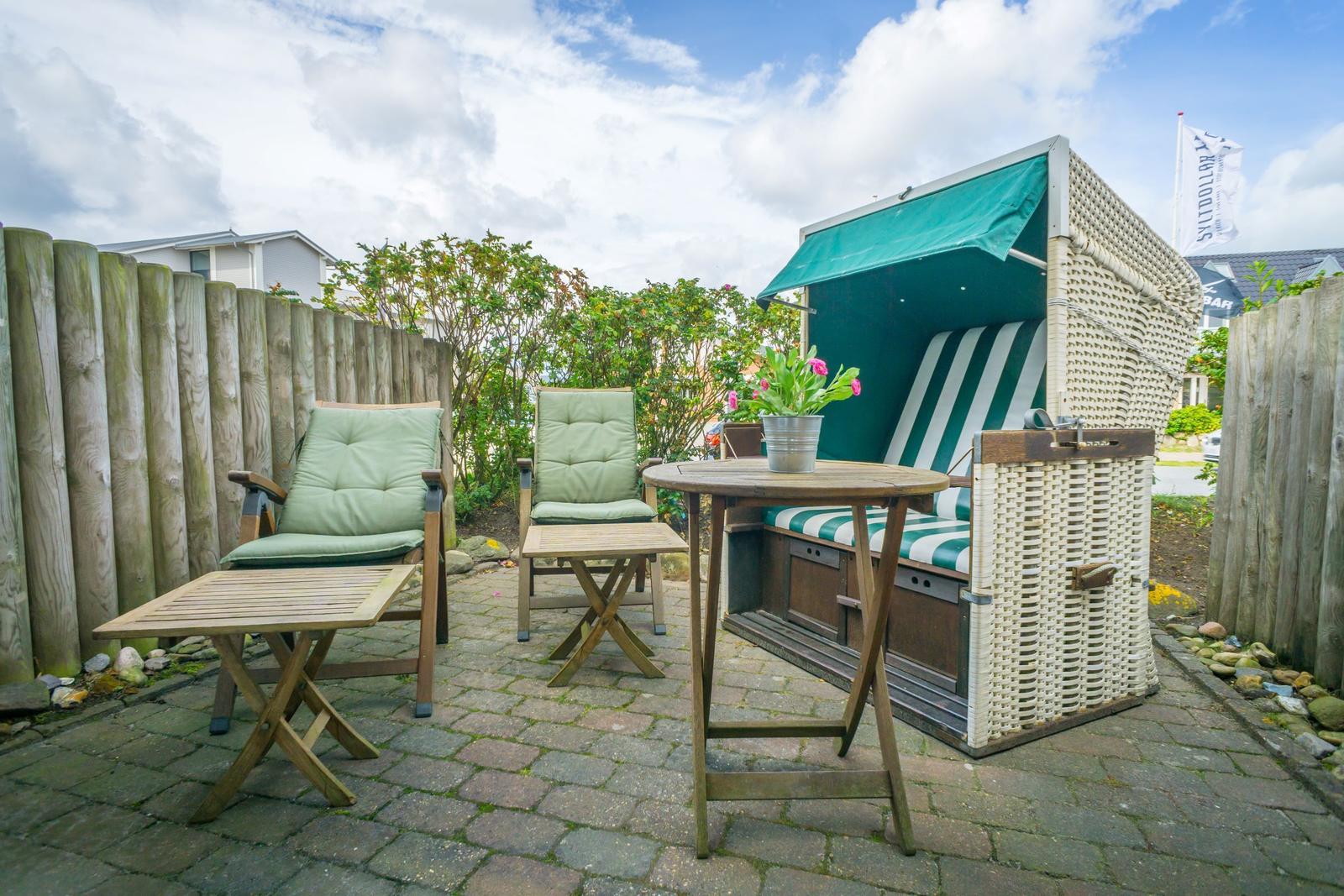 Terrasse mit Strandkorb, Tisch und zwei Stühlen. Holzplatten und grüne Kissen.