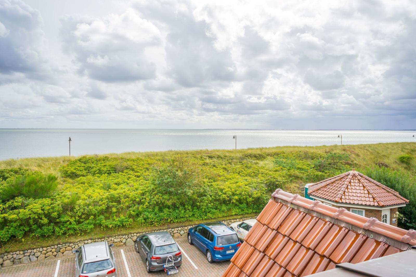 Vom Balkon aus Blick auf den Strand und das Meer mit grünen Dünen und geparkten Autos.