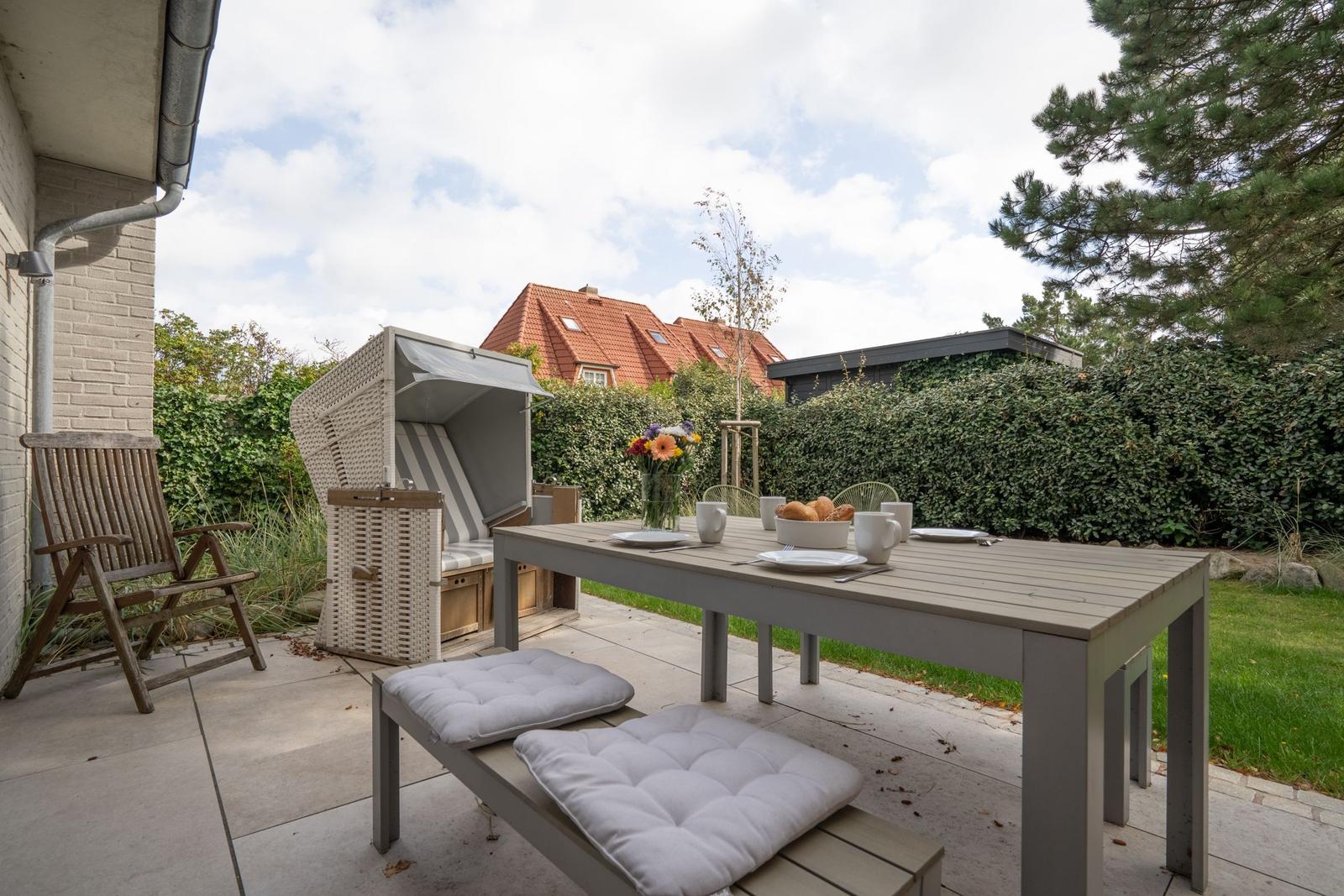 Terrasse mit Tisch, Stühlen und Strandkorb. Hinterhof mit Hecke und Bäumen.