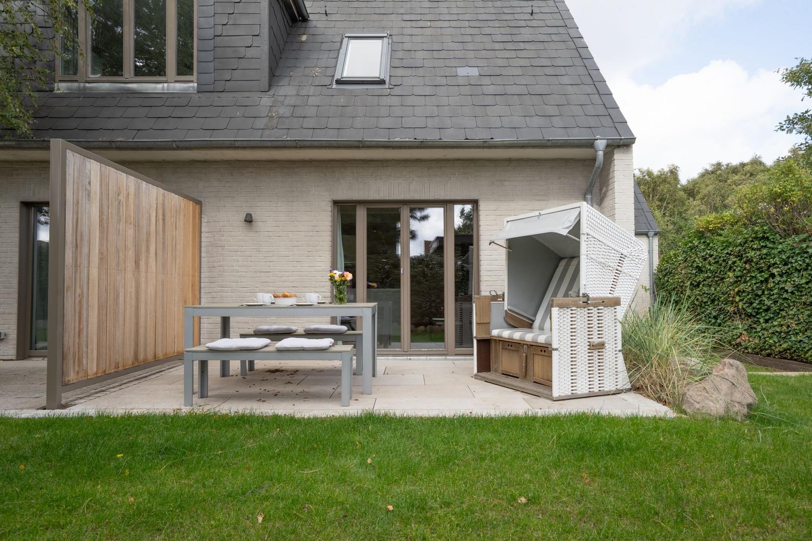 Terrasse mit Tisch, Bänken und Liegestuhl. Hinterhof mit Rasen und Hecke.