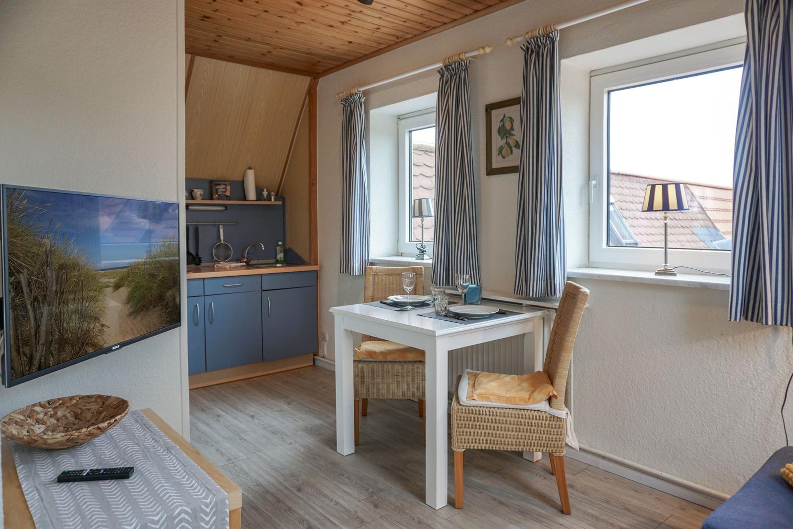 Wohnbereich mit Essplatz, TV und Küchenzeile. Fenster mit Vorhängen.