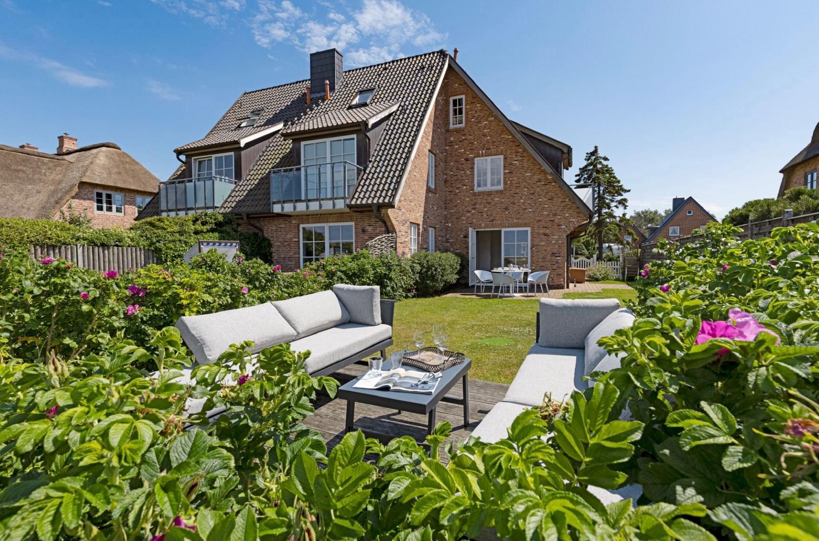 House with garden, terrace, and seating. Rose bushes and green plants surround the outdoor area.