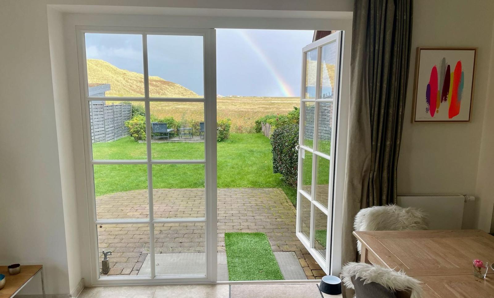 Through the glass door, one sees a paved courtyard with lawn and a rainbow over the dunes.