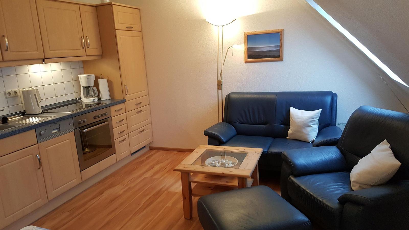 Living area with kitchen, sofa, and armchairs under sloped ceiling.