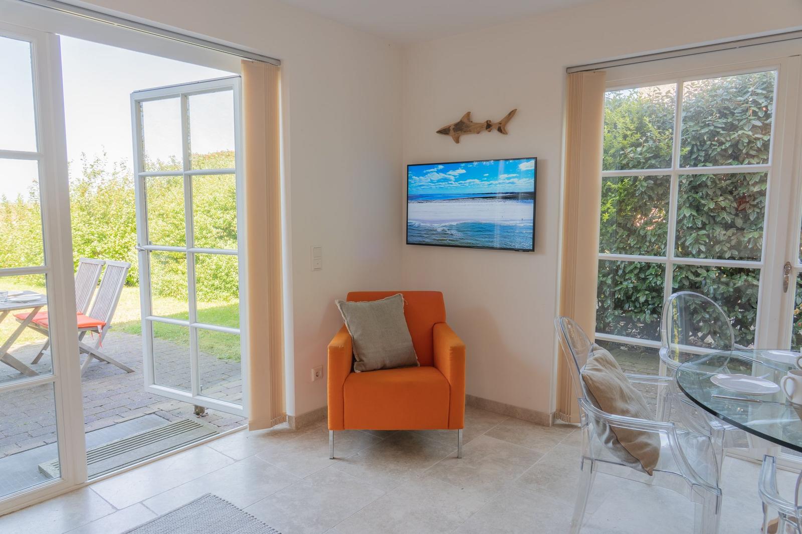 Ein Raum mit orangefarbenem Sessel, Fernseher und Glassitzen. Durch Glastüren ist eine Terrasse sichtbar.