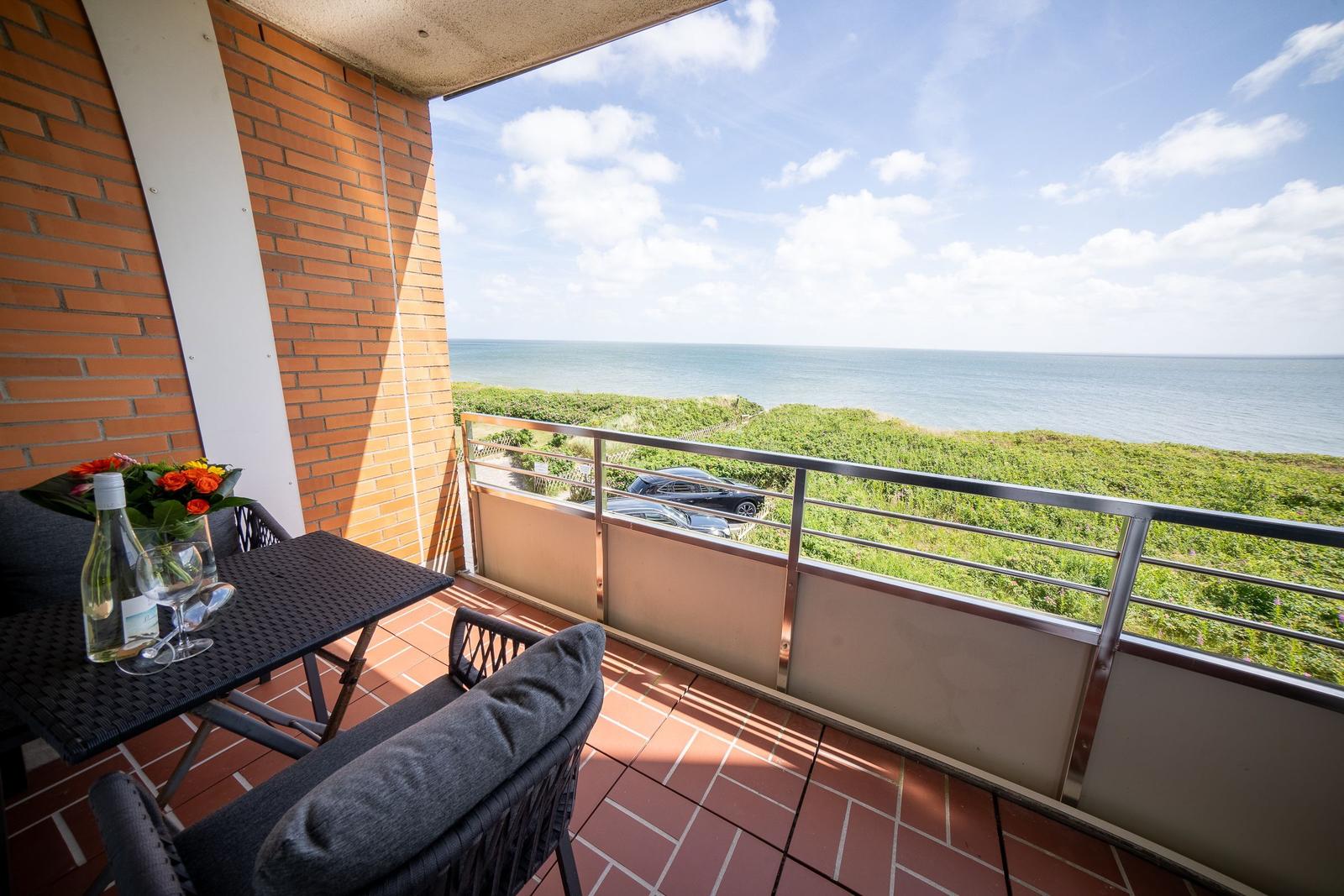 Terrace with table, chairs, and sea view. Behind the railing are grassy areas and a car visible.