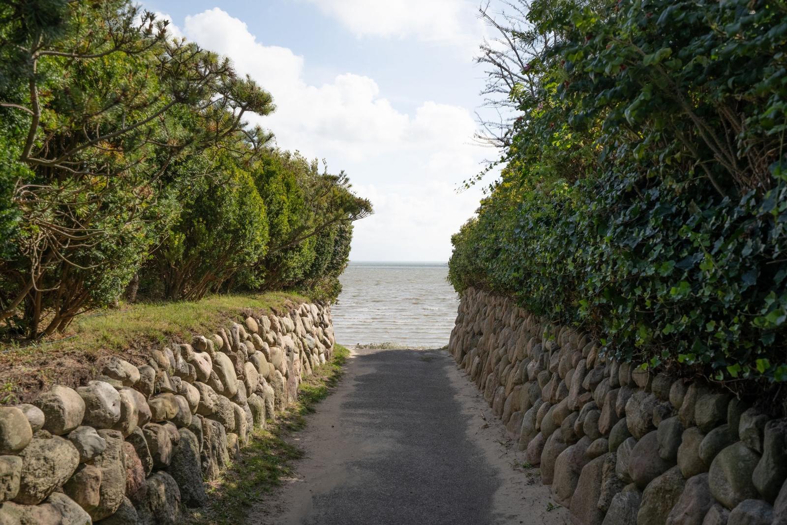 Steinmauerweg führt zum Meer. Grüne Bäume und Sträucher säumen den Pfad.