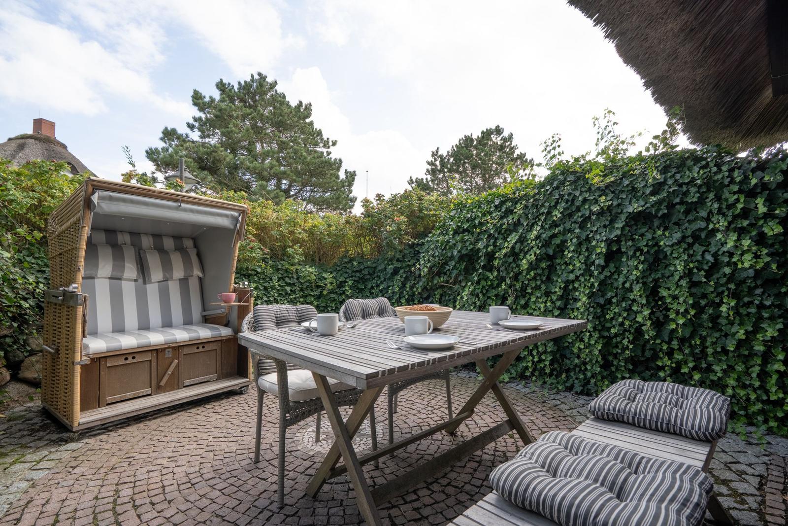 Terrasse mit Holztisch, Stühlen und Strandkorb. Hintergrund: grüne Hecke und Bäume.