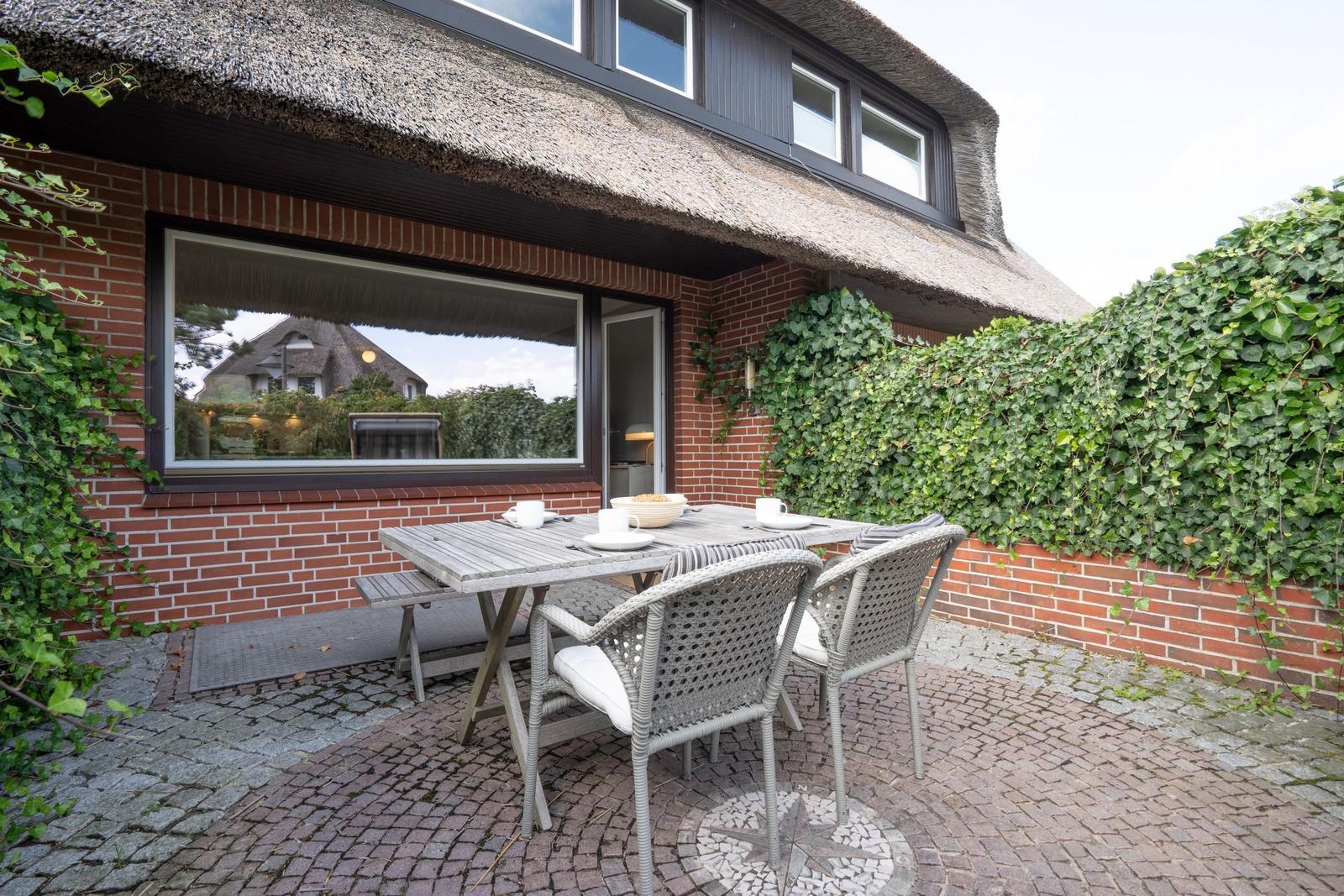 Terrasse mit Tisch und Stühlen vor einem Haus mit Schilfdecke und Kletterpflanzen.