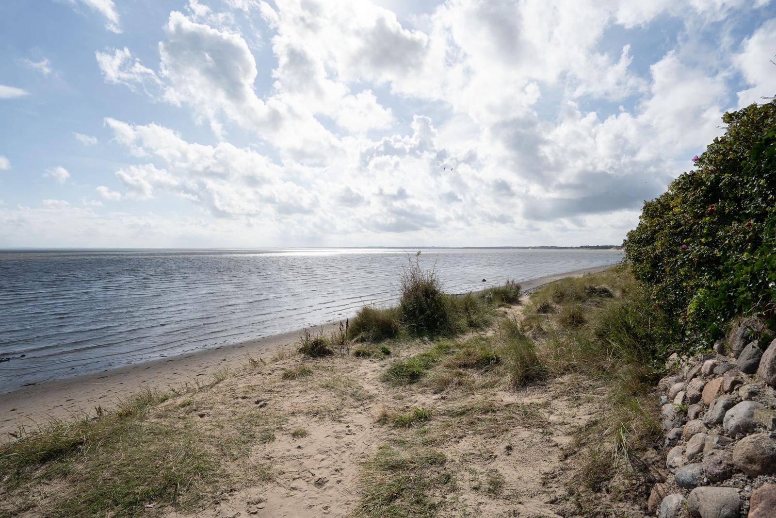 Strand mit Dünen, Gras und Steinmauer unter bewölktem Himmel.