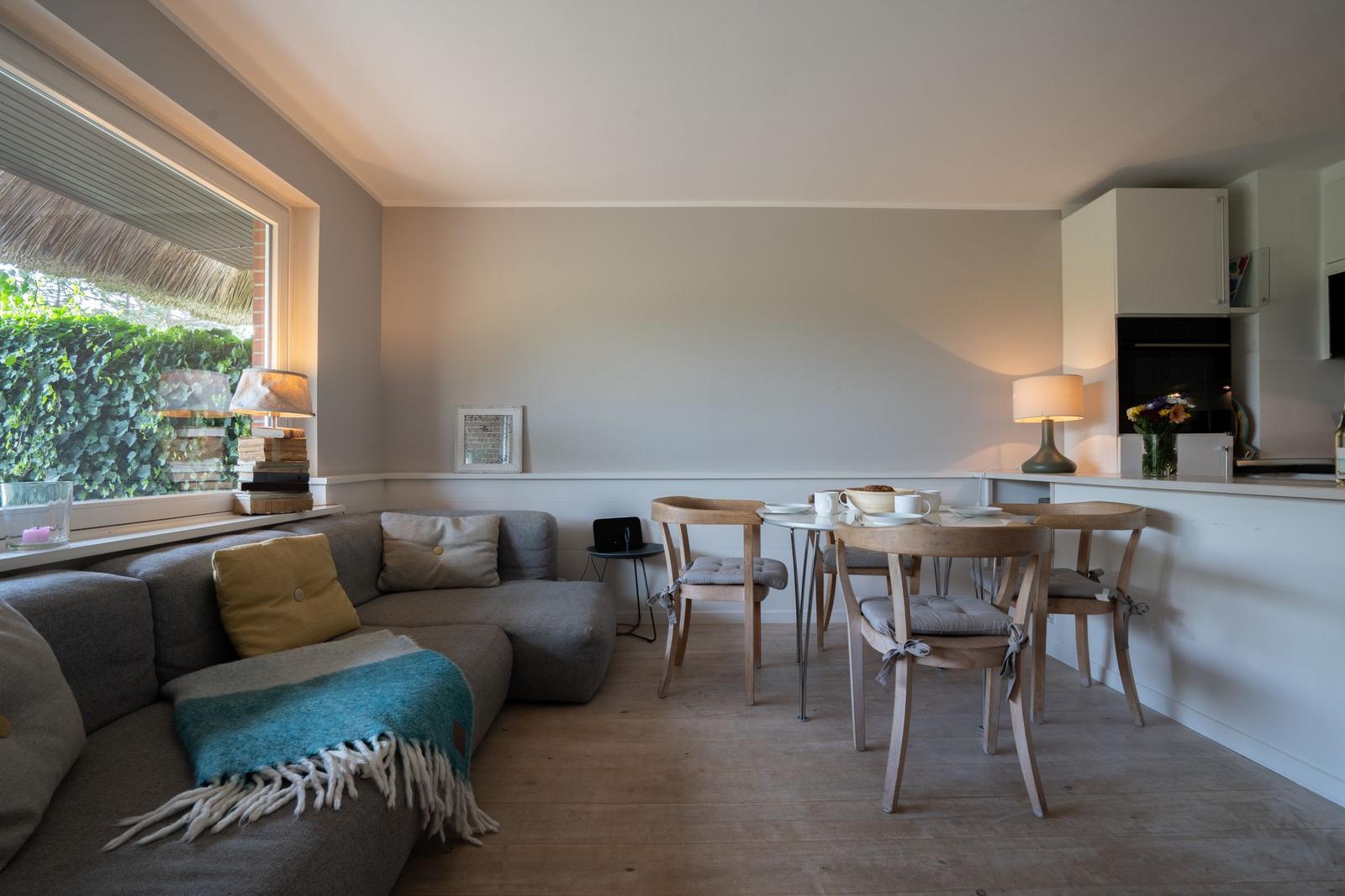 Wohnbereich mit Sofa, Esstisch und Küchenzeile. Fenster mit Blick auf grüne Hecke.