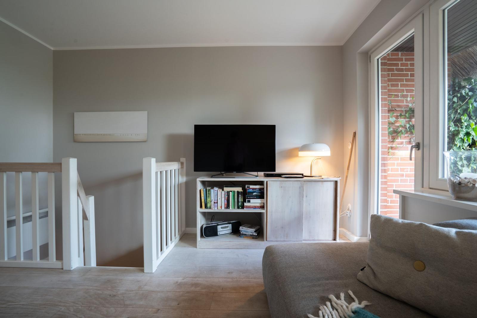 Wohnzimmer mit Sofa, Fernseher und Bücherregal. Fensterblick auf Außenbereich.