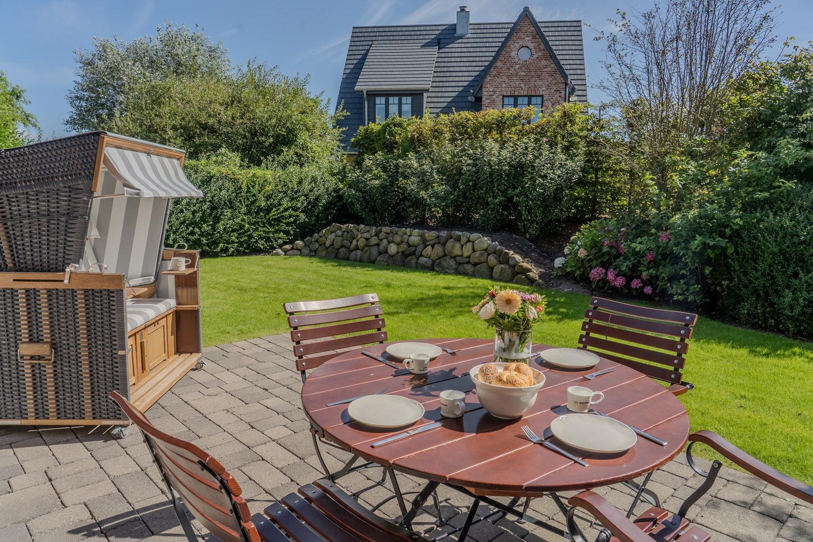 Runder Tisch mit Stühlen und Picknickdecke im Garten. Hintergrund: Haus und Grünflächen.