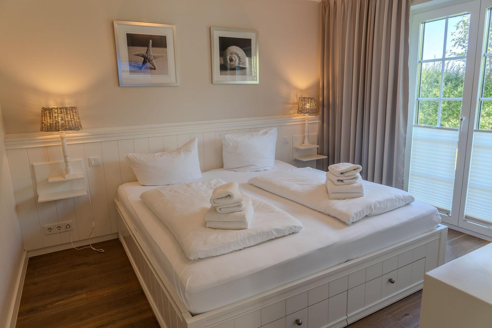 Schlafzimmer mit weißem Bett, zwei Nachttischen und Fenster mit Blick auf Grün.