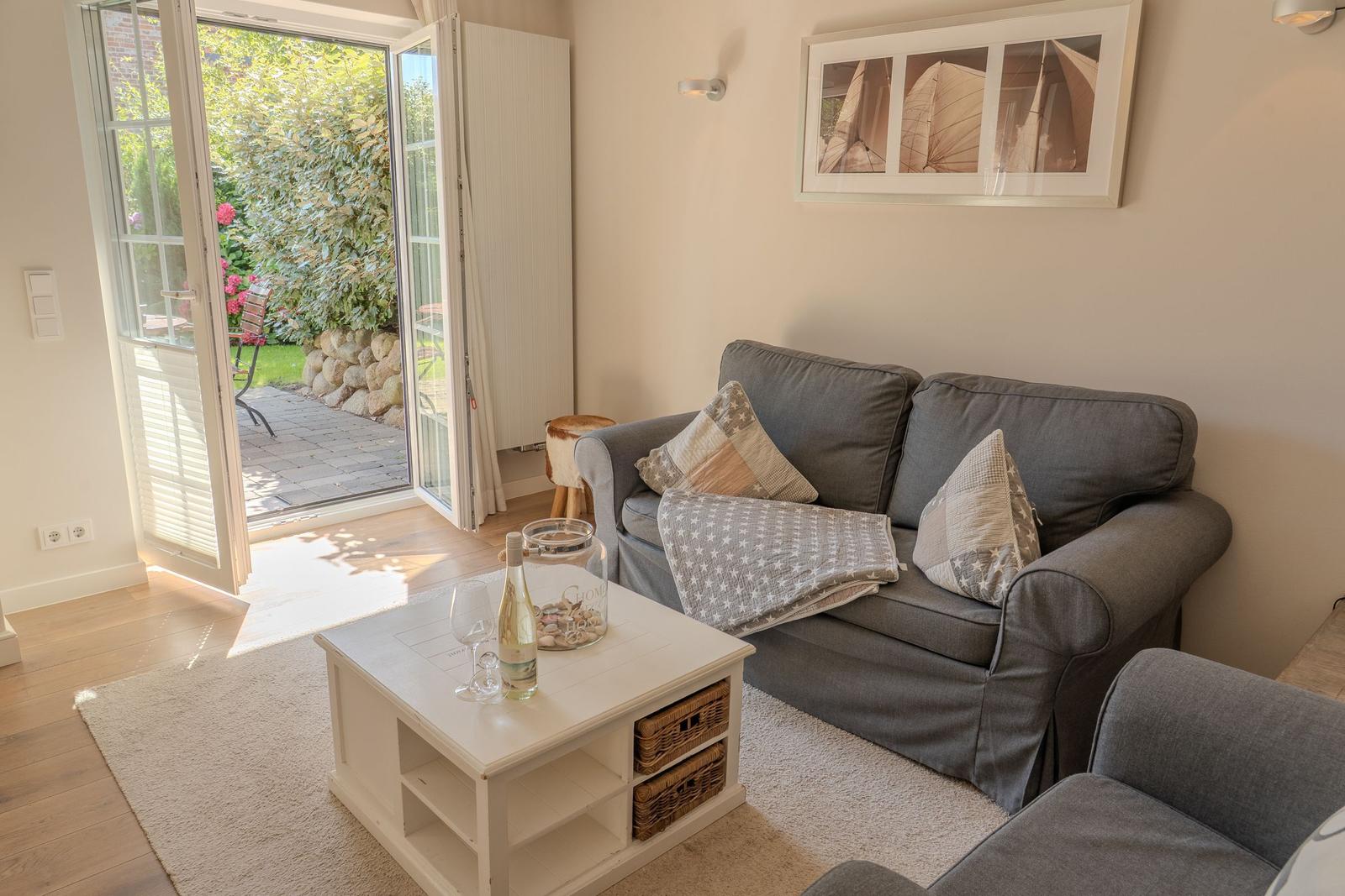 Grauer Sofa mit Kissen und Decke, weißer Couchtisch, Terrasse mit Gartenblick.