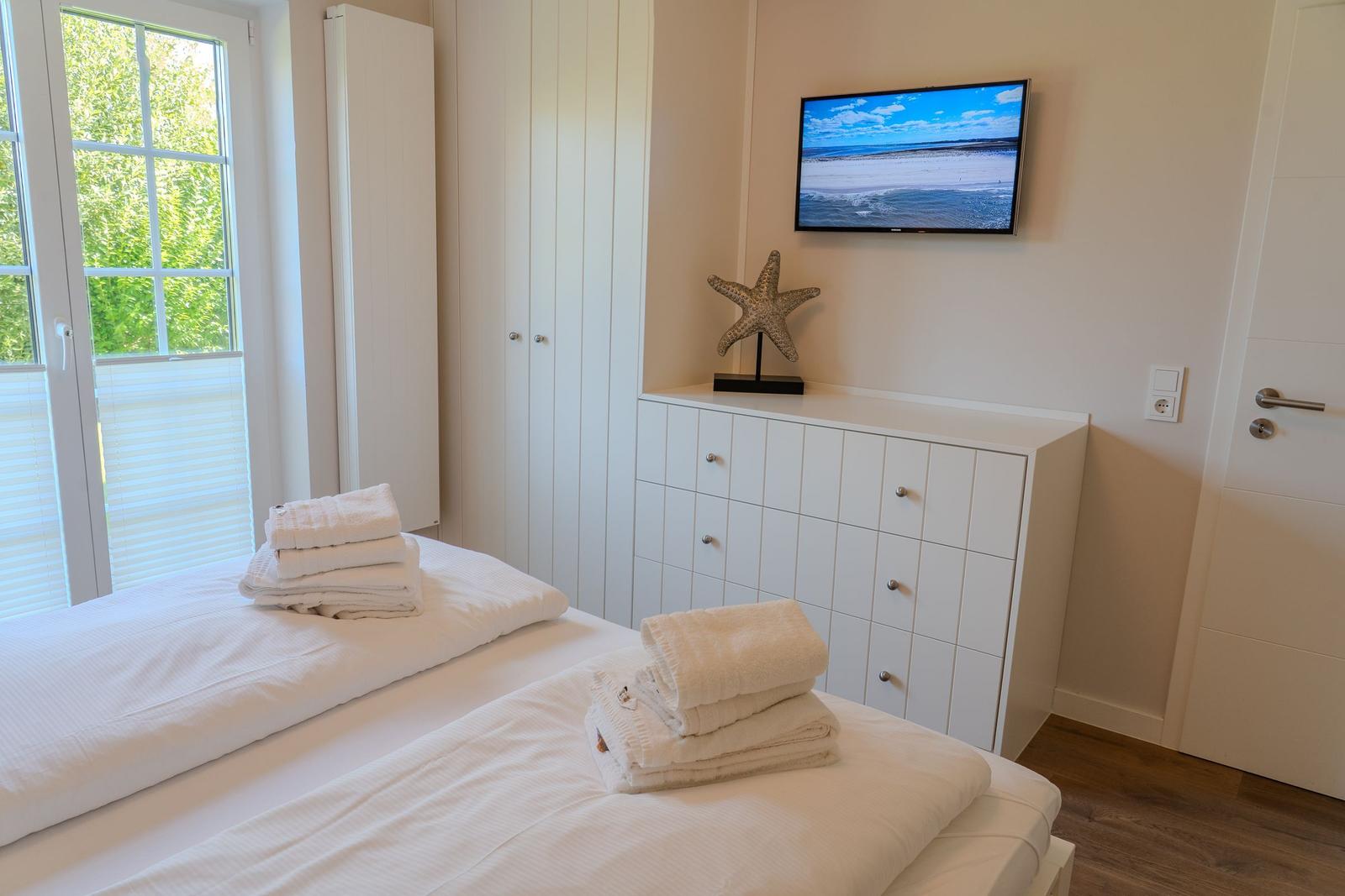Schlafzimmer mit Bett, TV und Garderobe. Fenster mit Blick auf Grün.