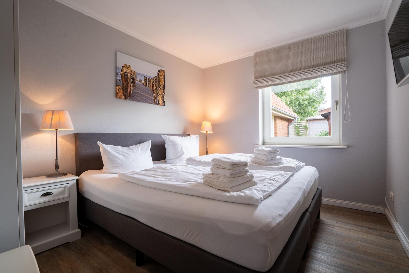 Schlafzimmer mit Bett, Nachttisch und Fenster. Auf dem Bett liegen gefaltete Handtücher.