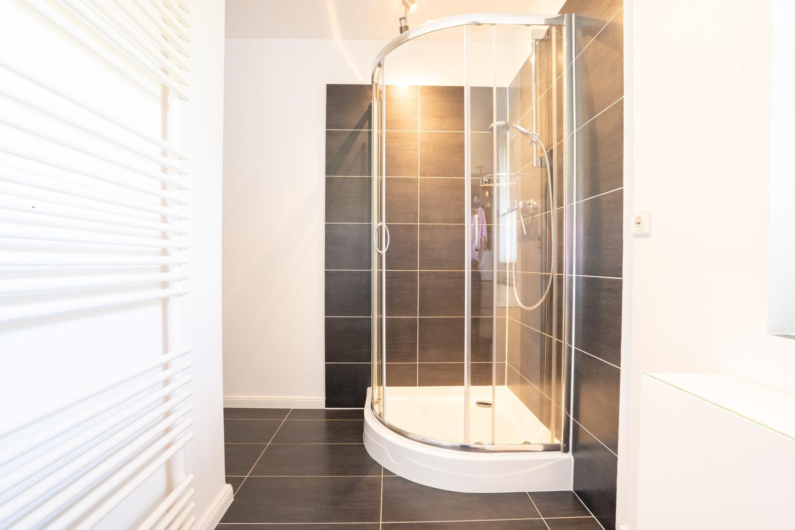 Dusche mit Glaswand und dunklen Fliesen. Heizung links. Bodenfliesen schwarz.