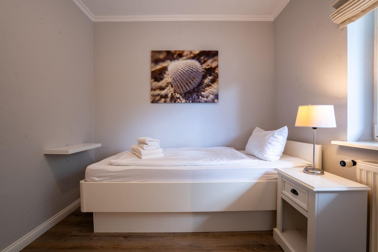 Schlafzimmer mit weißem Bett, Nachttisch und Wandbild mit Muschel.