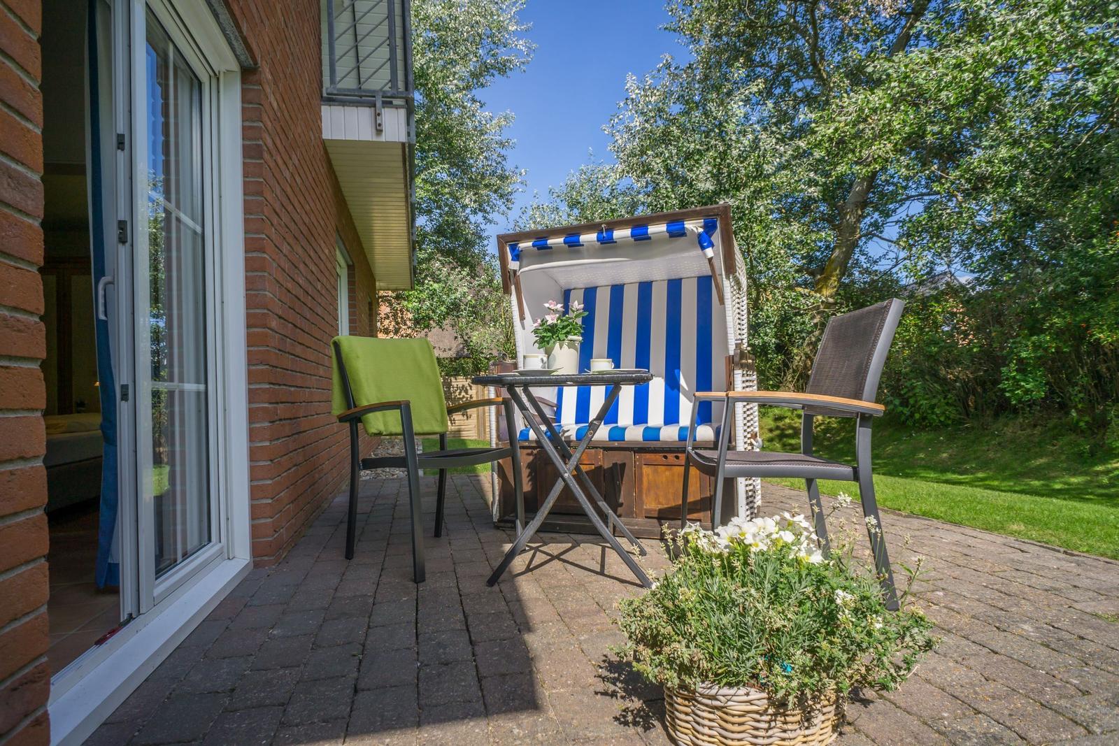 Terrasse mit Strandkorb, Tisch und Stühlen. Hintergrund: Grünfläche und Bäume.