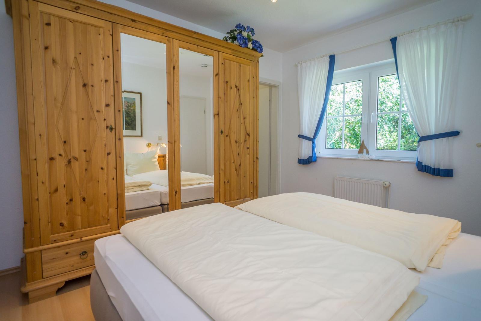 Schlafzimmer mit Doppelbett, Holzschrank und Fenster mit weißen Vorhängen.