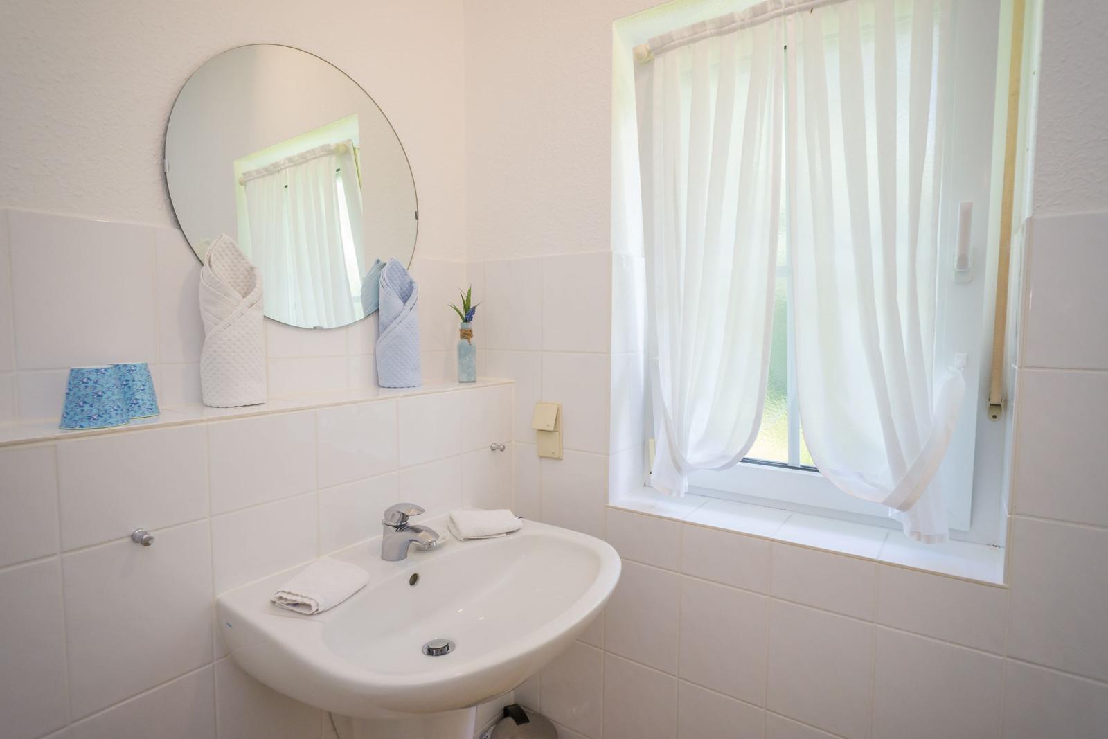 Bad mit Waschbecken, Spiegel und Fenster mit weißen Vorhängen.