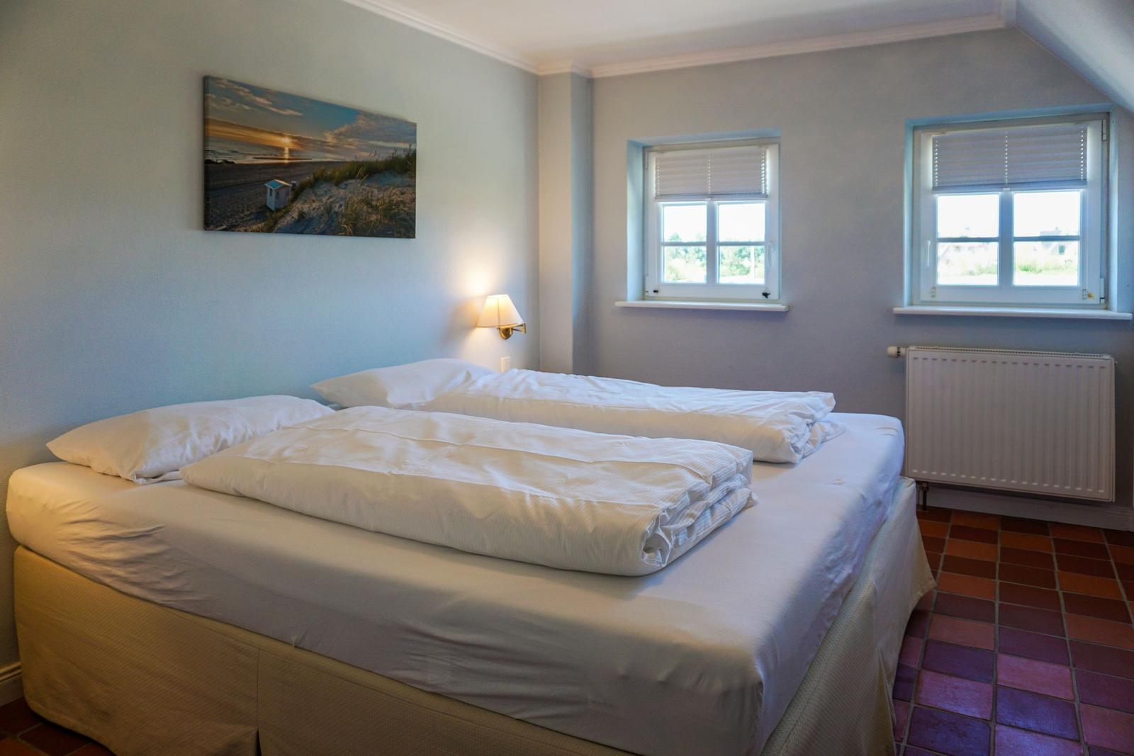 Schlafzimmer mit Doppelbett, zwei Fenstern und Wandbild.