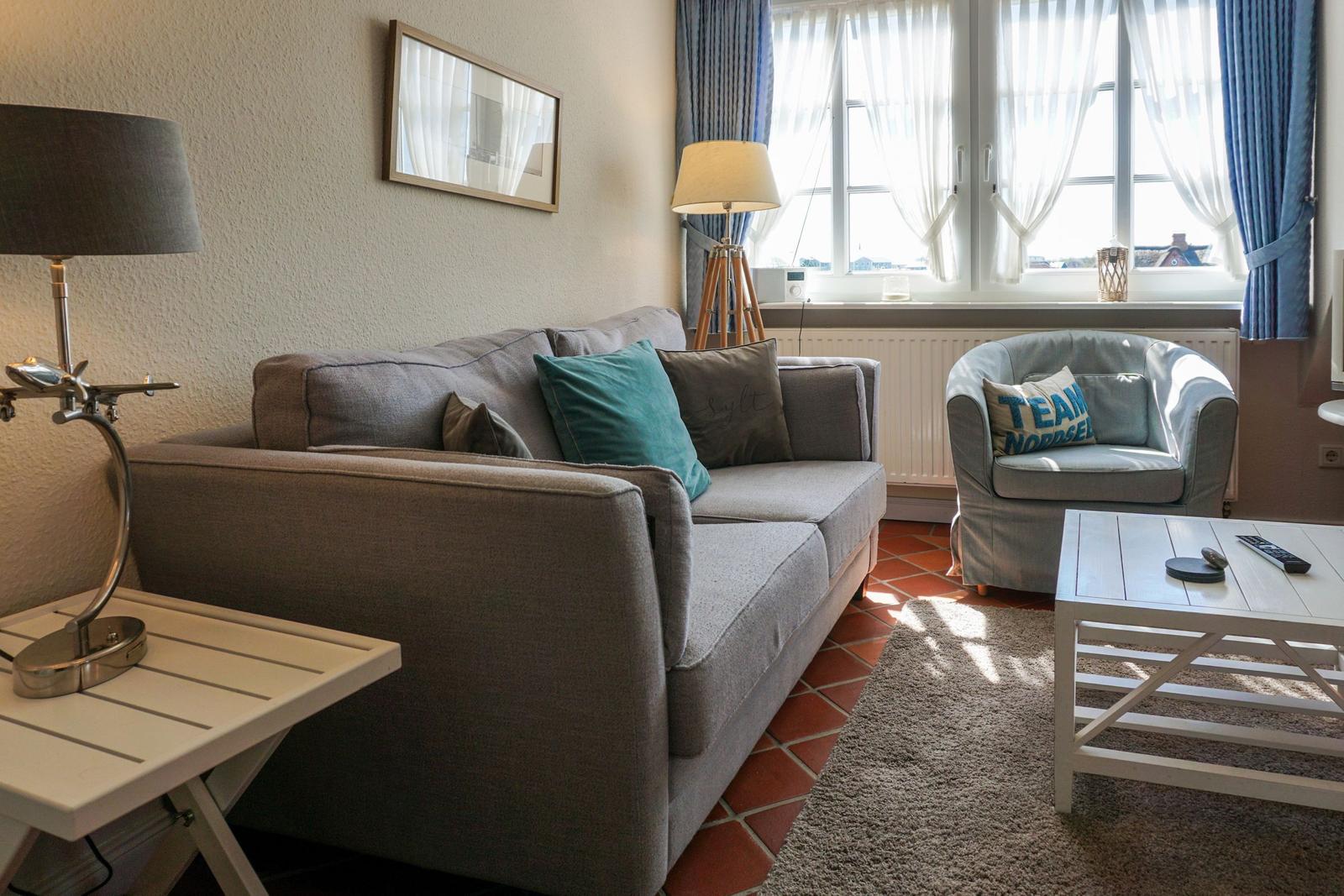 Wohnzimmer mit Sofa, Sessel und Tisch. Fenster mit Vorhängen. Teppich und Bodenfliesen.