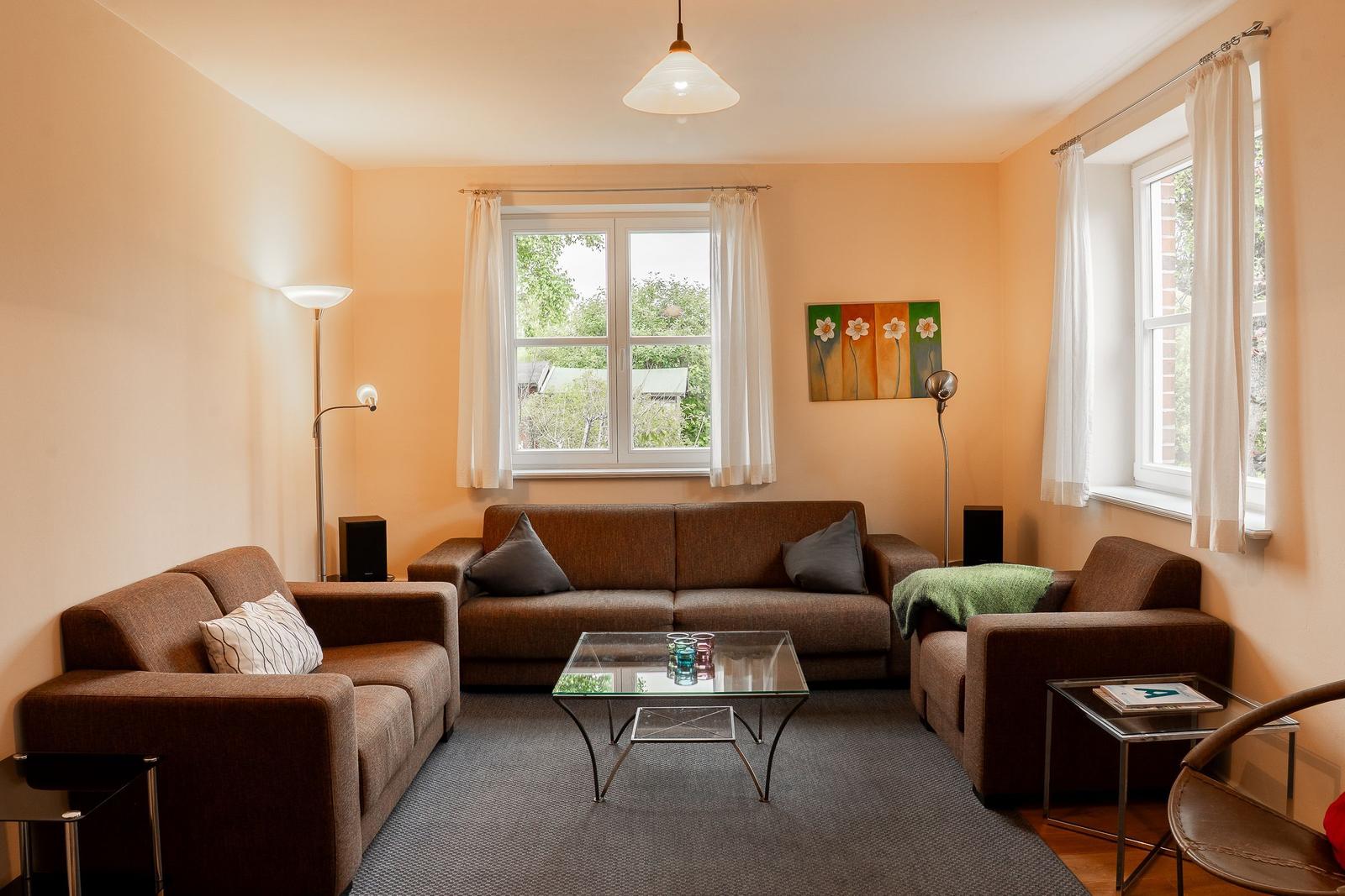 Living room with sofa, armchairs, and glass table. Windows with curtains. Wall art with flowers.