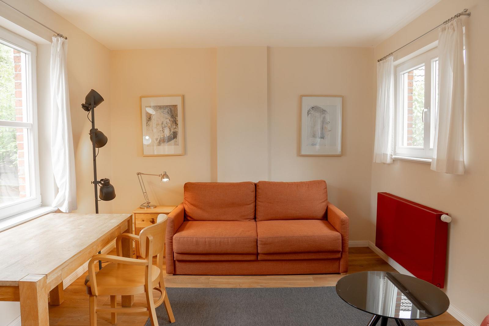 A room with an orange sofa, wooden table, chair, and two windows.