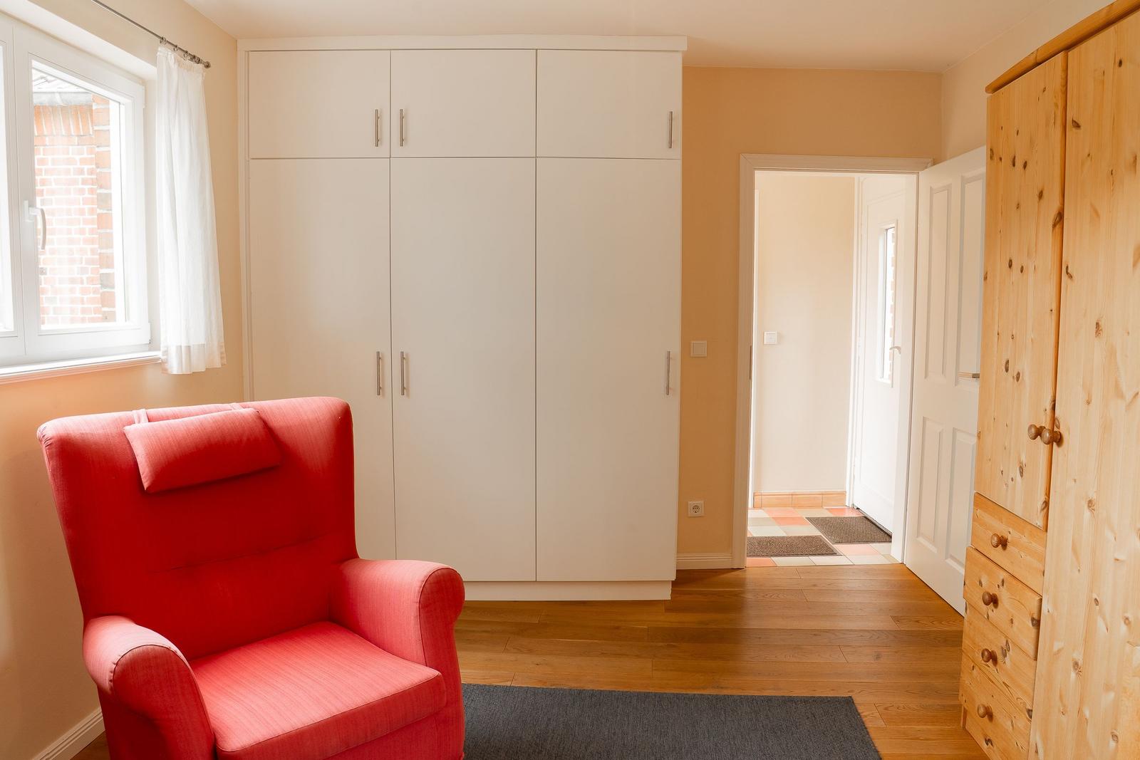 A room with a red armchair, white wardrobes, and wooden furniture.