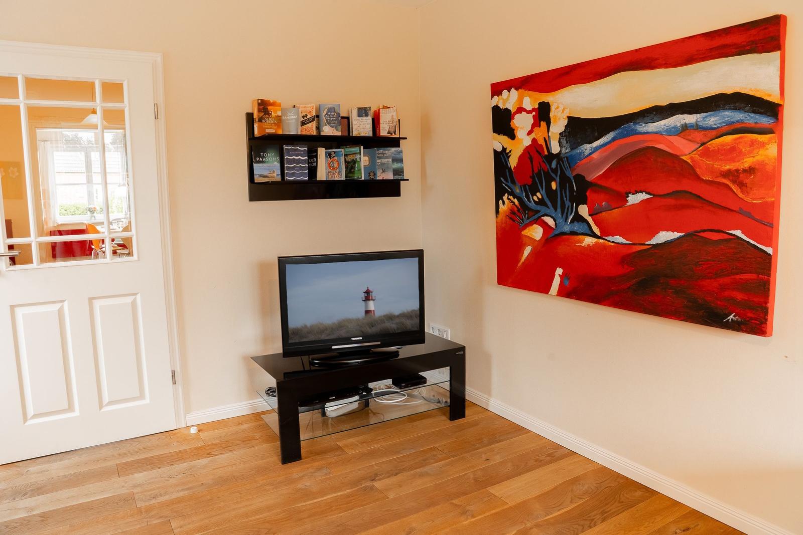 Living room with TV, wall art, and bookshelf. Wooden floor and door to kitchen.