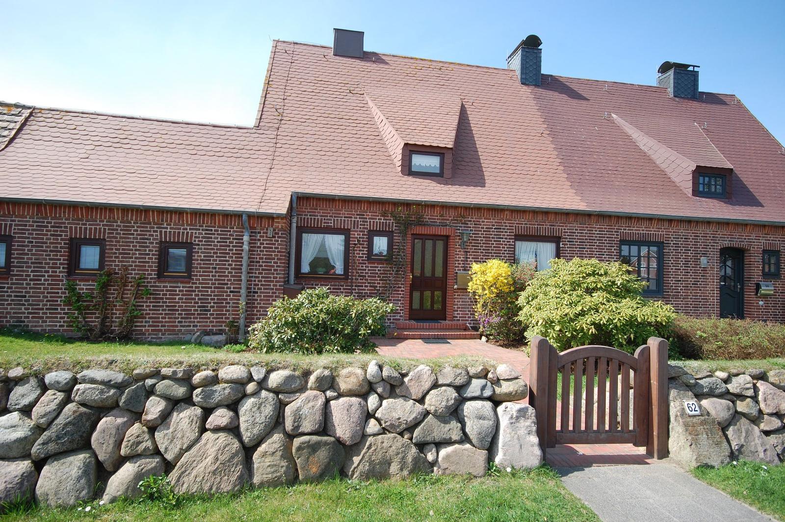 Red tile roof, brick house with entrance door and stone wall. Wooden gate with number 62.
