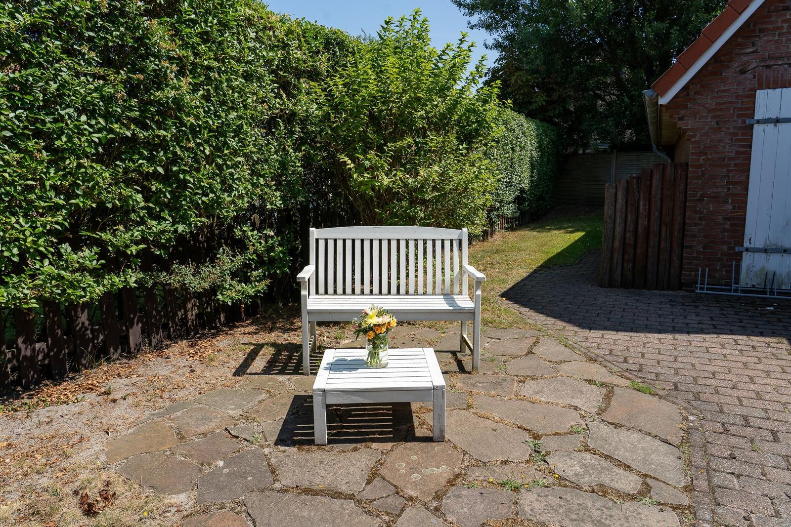 Weiße Bank und Tisch mit Blumen auf Steinplatten im Garten.