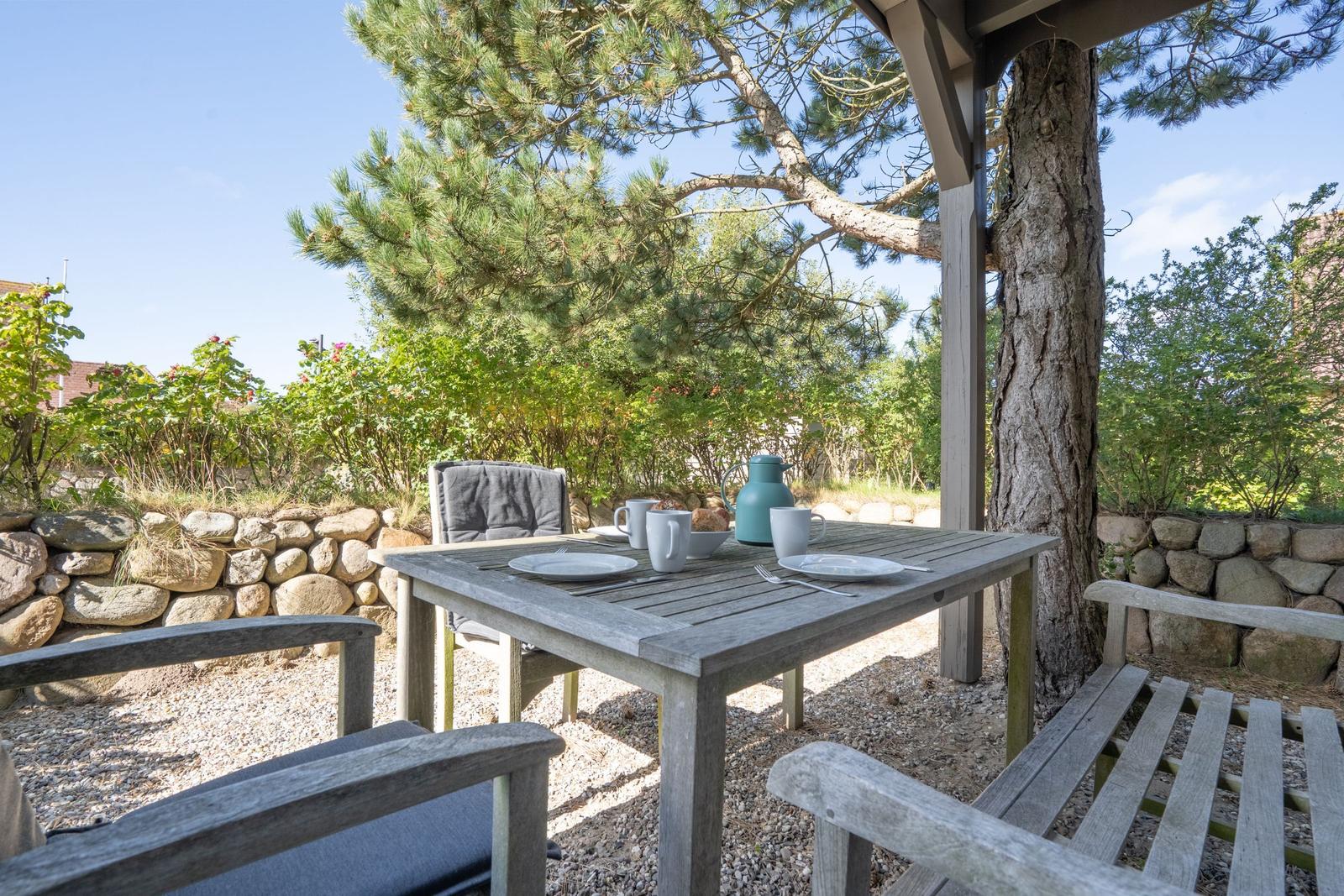 Holztisch mit Stühlen unter einem Pavillon, umgeben von Bäumen und Steinmauern.