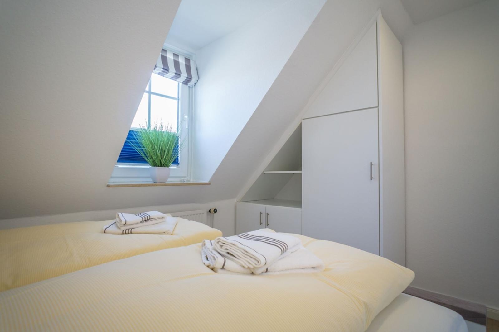 Bedroom with sloped ceiling, bed, and wardrobe. Window with plant and striped roller blind.