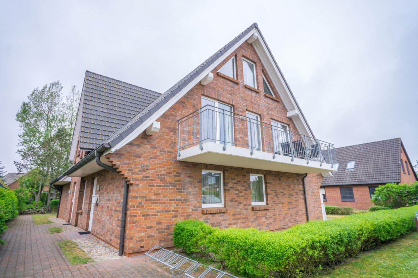 Zweistöckiges Haus mit Balkon und rotem Backsteinbau. Grünflächen und gepflasterte Wege.