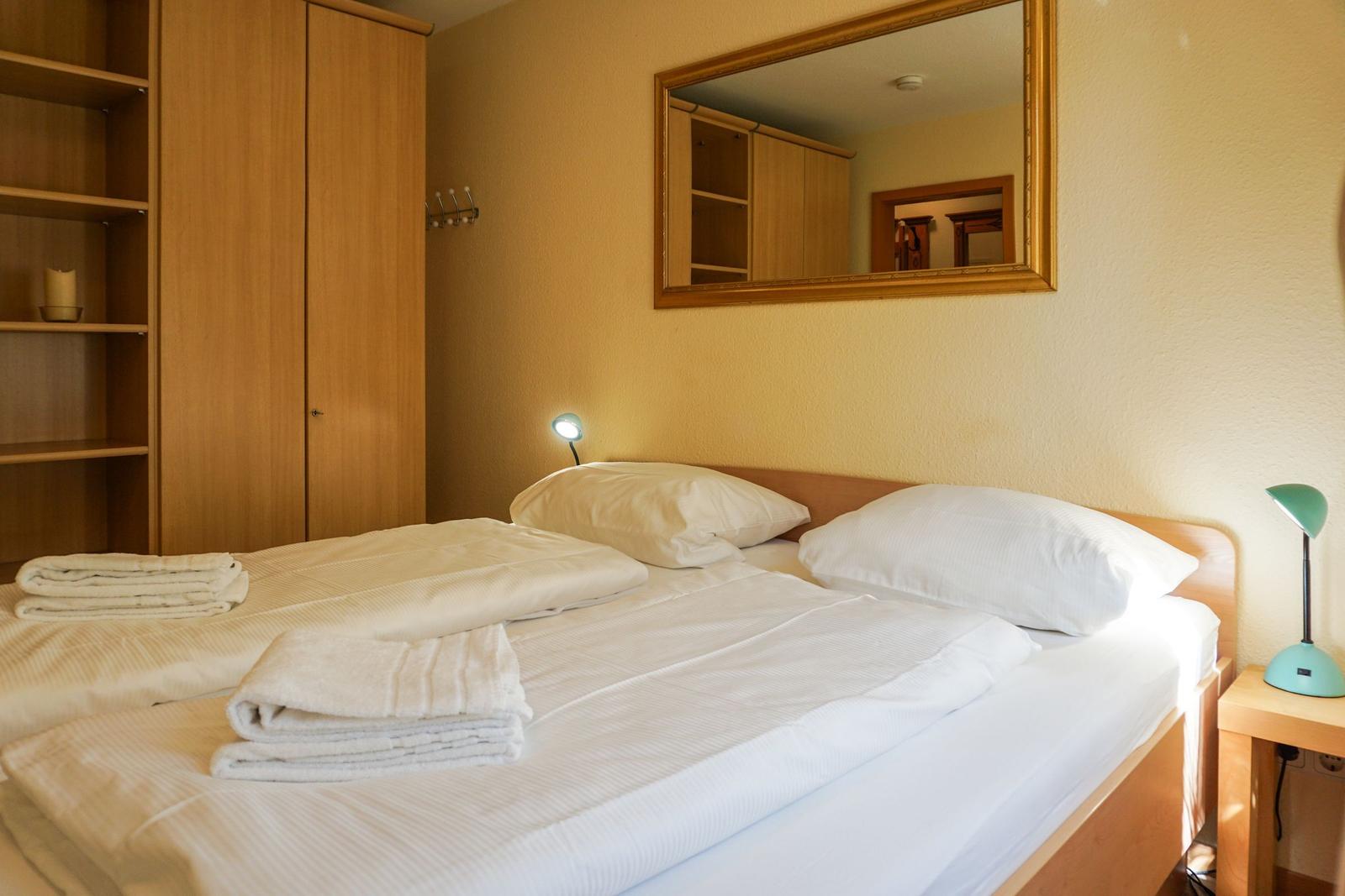 Doppelbett mit weißen Laken, Holzkommode und Spiegel an der Wand.