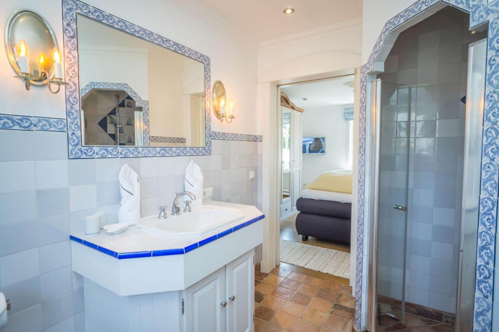 Badezimmer mit Waschbecken, Spiegel und Dusche. Blick in Schlafzimmer mit Bett.