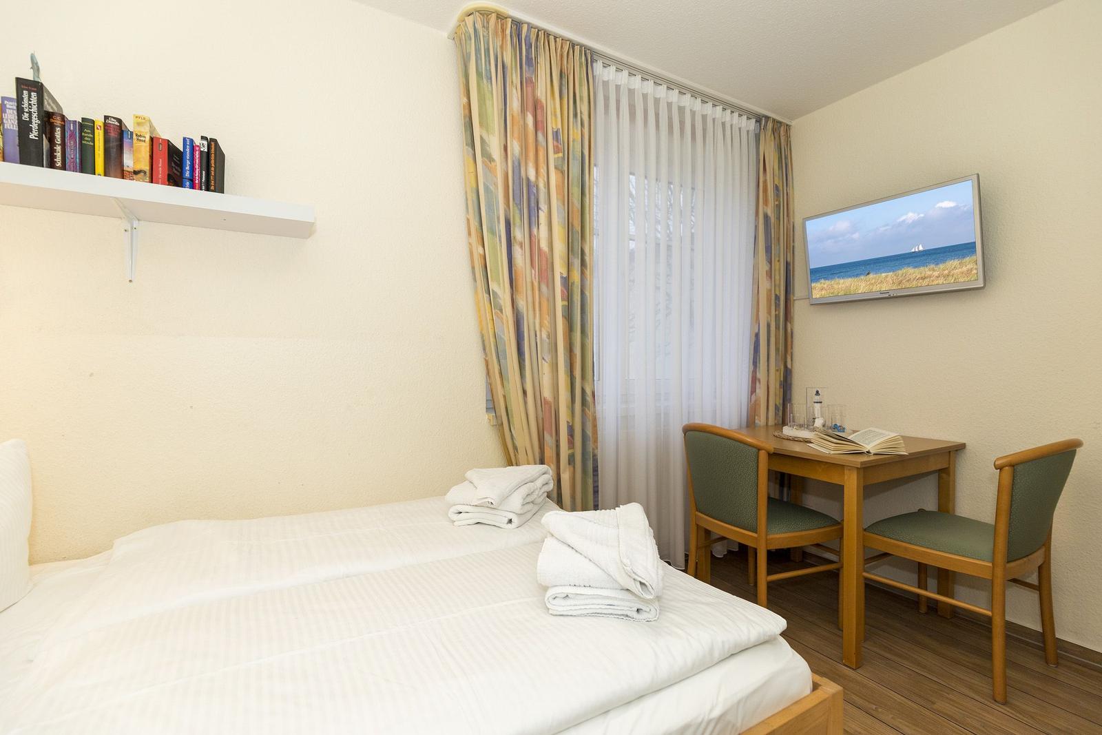Zimmer mit Bett, Schreibtisch, TV und Bücherregal. Fenster mit Vorhängen.