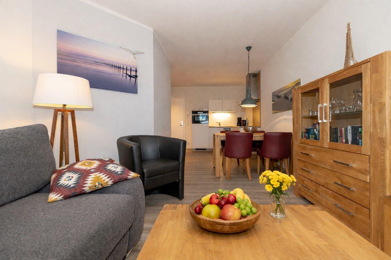 Living area with sofa, armchair, and dining area. Wooden table, fruit bowl, and bookcase visible.