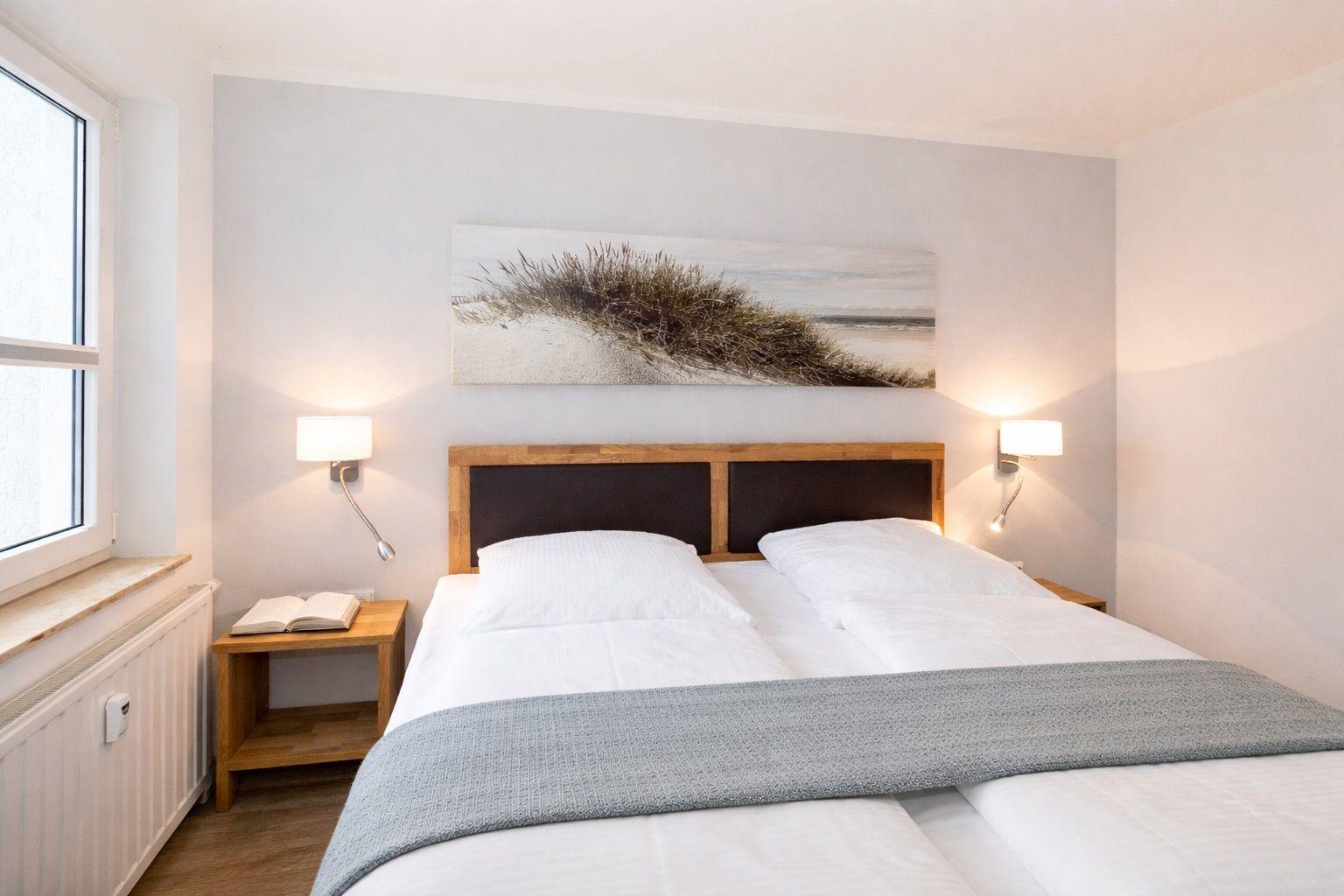 Bedroom with double bed, wooden side table, and wall art. Two wall lamps and window with radiator.