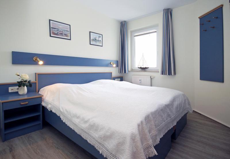 Bedroom with blue bed, nightstand, and window with sailboat. Wall pictures and curtains.