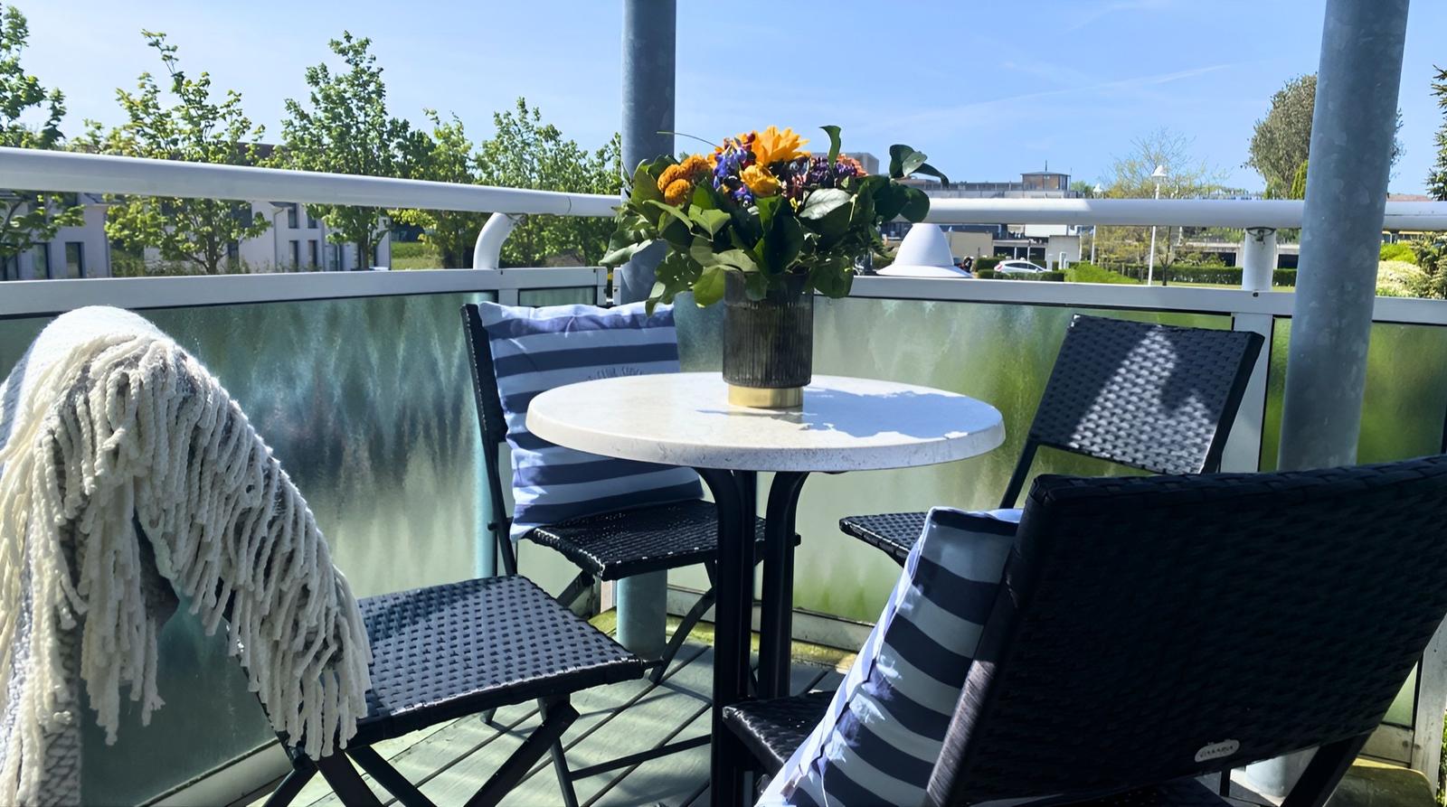 Terrasse mit Tisch, Stühlen und Blumenstrauß. Glasgeländer und Blick auf Grünflächen.