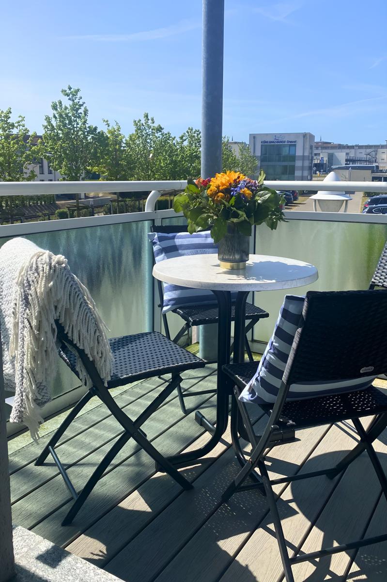 Terrasse mit Tisch, Stühlen und Blumenarrangement. Holzdielen und Glasgeländer.