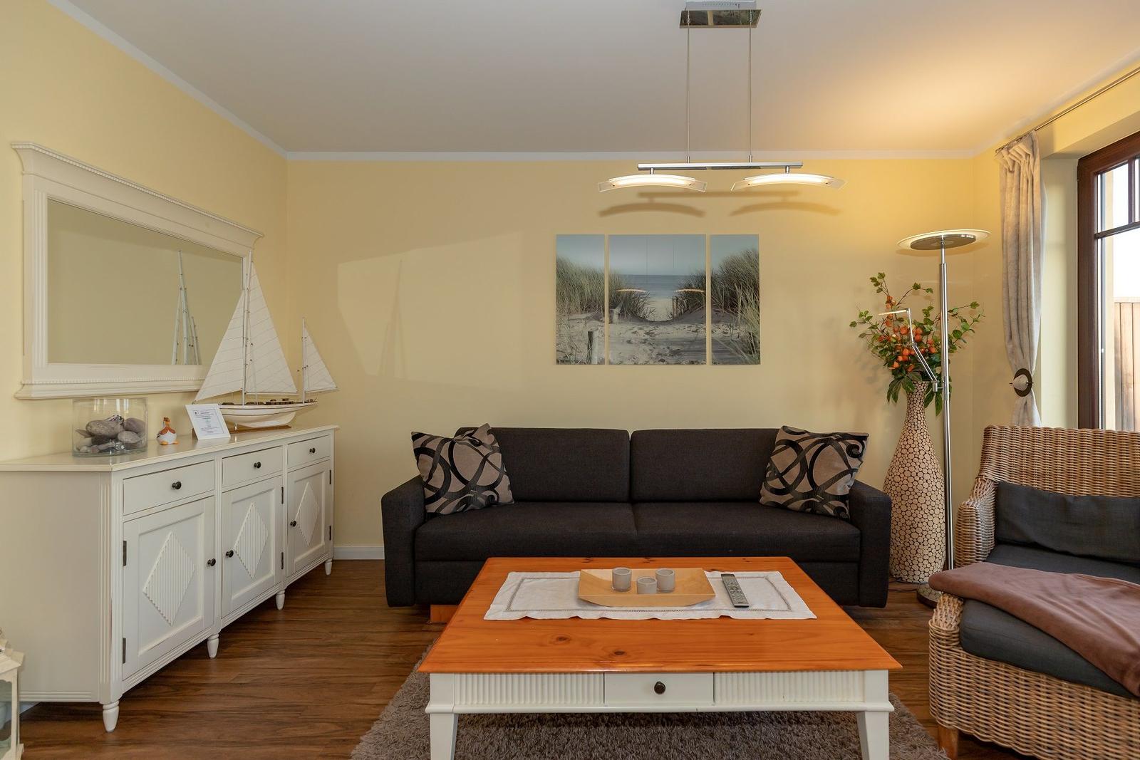 Ein Wohnzimmer mit Sofa, Couchtisch und Wandbild. Links steht ein Sideboard mit Segelbootmodell.