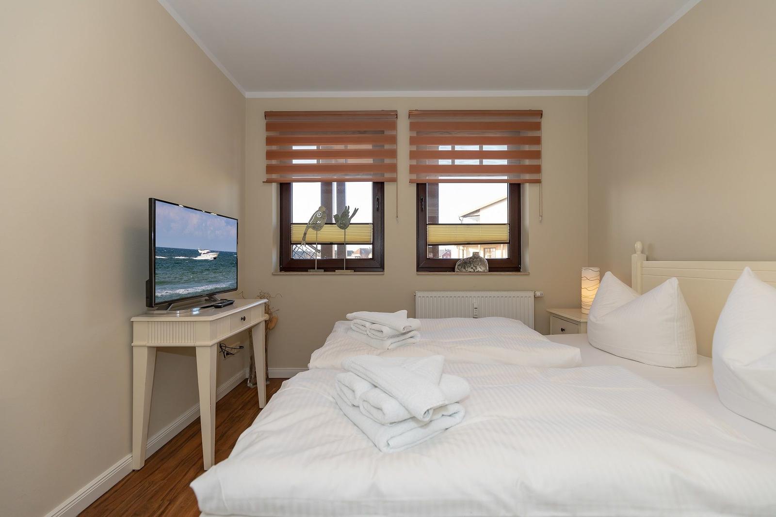 Schlafzimmer mit Bett, Fernseher und zwei Fenstern mit Jalousien.
