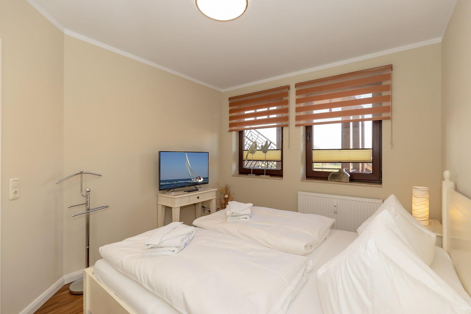 Schlafzimmer mit Doppelbett, TV und zwei Fenstern mit Jalousien.