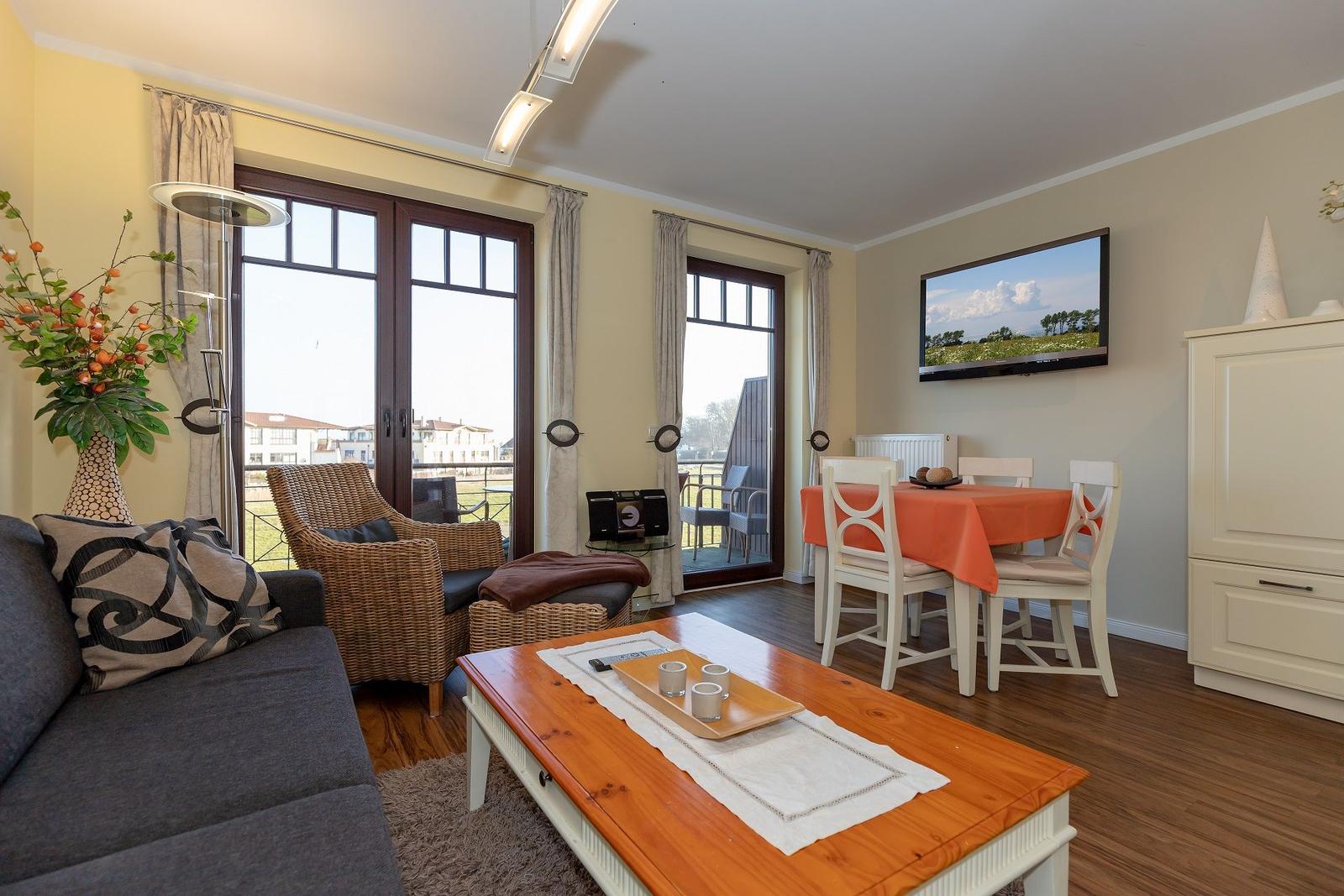 Wohn- und Essbereich mit Sofa, Esstisch und TV. Große Fenster mit Blick auf Außenbereich.