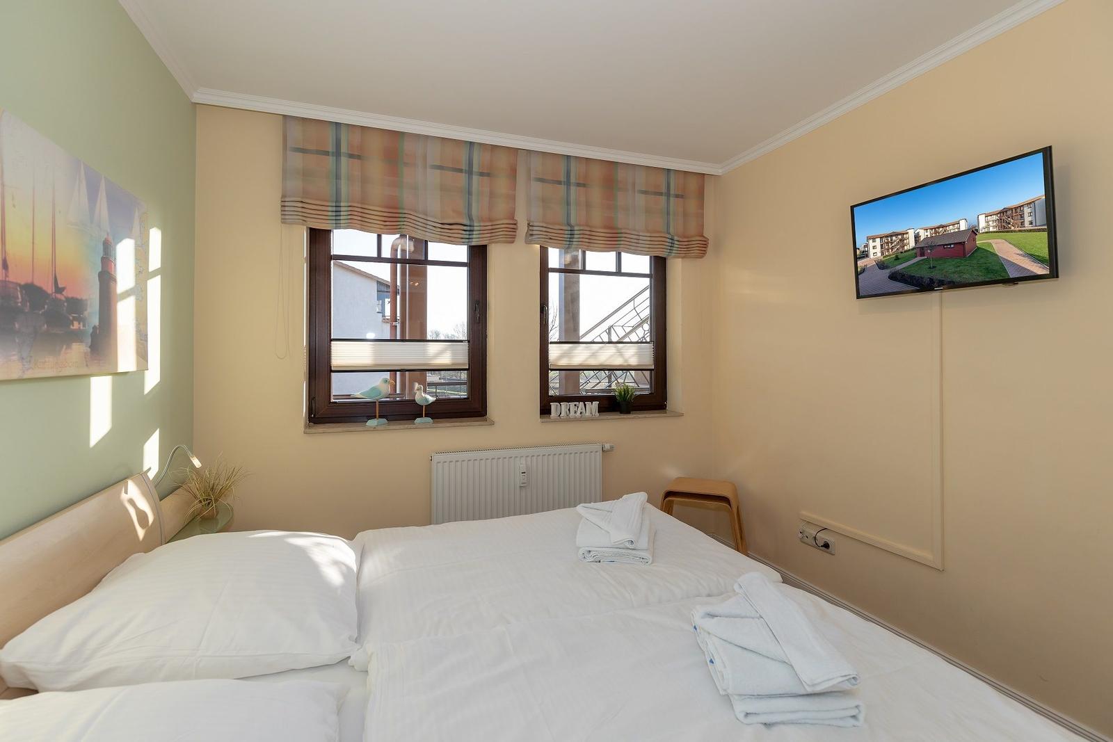 Schlafzimmer mit Doppelbett, Fenster, Fernseher und Wandbild.
